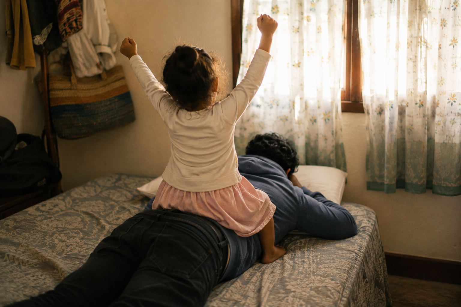 Dans une chambre chaleureuse, un homme allongé à plat ventre sur un lit sourit les yeux fermés pendant qu'une petite fille assise sur son dos joue au cheval les bras levés